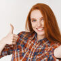 Beautiful redhead girl smiling happy and showing thumb-up while taking selfie on smartphone, front camera view, white background.