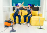 Satisfied couple of cleaners in blue uniforms giving each other high five and continuing to clean the floor in the kitchen.